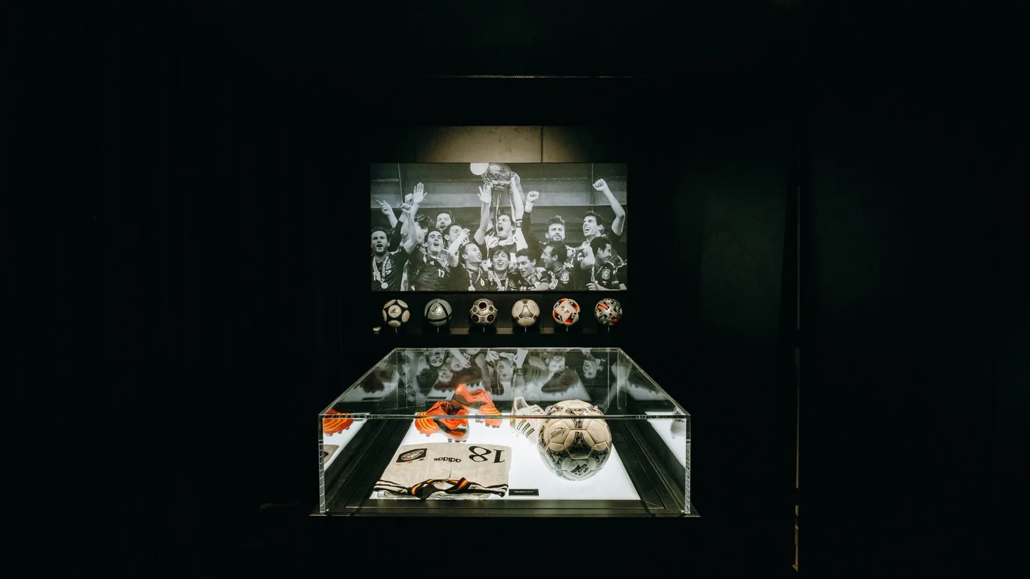 adidas-Ausstellung zur UEFA EURO 2024 – Acrylvitrine mit Trikot, Fußballschuhen und Spielball vor einer Reihe von EM-Bällen